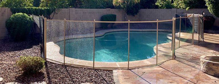 A backyard swimming pool in Arizona surrounded by a mesh pool fence for safety, with lush green bushes and trees in the background. The pool is clean and has a curved shape, and the pool fence is securely installed on the stone pavement around the pool area.