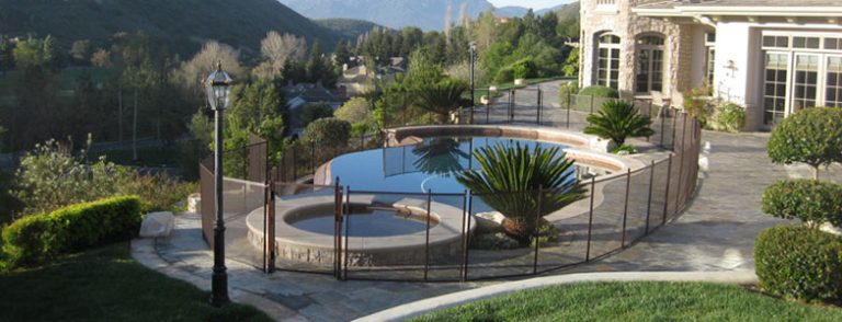 An elegant backyard pool in Ventura with a scenic view of mountains and lush greenery. The pool area is surrounded by a stylish mesh pool fence for safety, ensuring both security and aesthetic appeal. The landscape features well-maintained bushes, a lamppost, and a luxurious home in the background.
