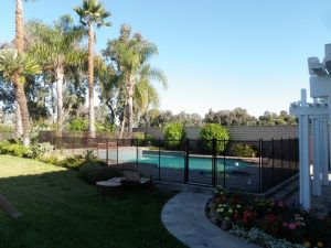 Backyard pool in Orange County with black mesh safety fence, lush greenery, a flower bed, lounge chair, and a brick wall in the background.