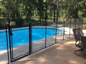 Black safety fence with a gate surrounding a pool, patio chairs nearby, and lush green trees in the background in Tulsa.