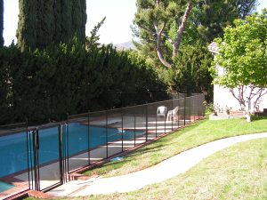 Inland Empire pool fence surrounding a backyard pool with tall privacy hedges, lawn chairs, and a garden path.