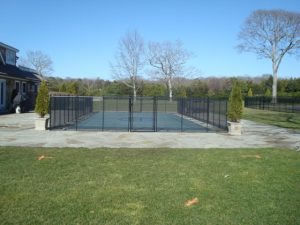 Pool fence encloses a rectangular pool with a stone deck, set in a spacious backyard with potted shrubs and leafless trees in Long Island.