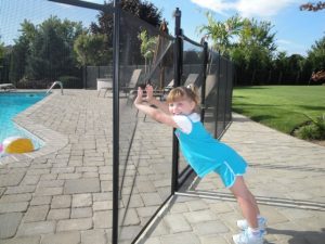 Young girl leans on a black mesh pool safety fence surrounding a pool with a patio and green lawn in the background.