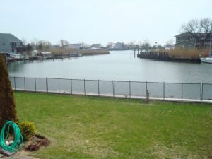 Mesh pool fence on a grassy lawn overlooking a calm waterway with docks and boats, set against a backdrop of waterfront homes on Long Island.