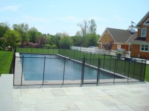 Black mesh pool fence around a rectangular pool in a spacious backyard with a paved patio and house.