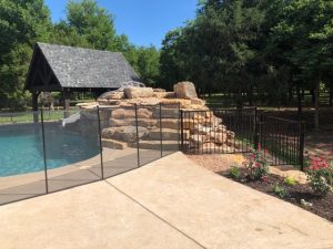 Pool fence installation in Tulsa featuring a black safety fence around a pool with a rock waterfall and a garden area with flowers.