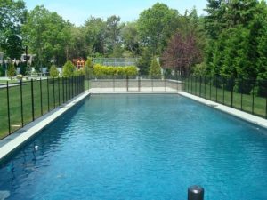 Pool fence surrounding a rectangular pool in a spacious Long Island backyard with lush greenery and a play area in the background.