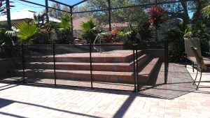 Black pool fence surrounding an outdoor spa with brick steps, lush plants, and patio furniture in a screened-in area.