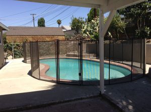 Backyard pool in Los Angeles with a black mesh safety fence, red brick pool edging, surrounded by greenery, and a patio area.