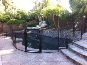Black mesh pool fence in Los Angeles surrounding a pool with a waterfall feature, lush greenery, and stone patio steps in the background.