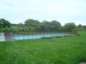 Secure pool fence surrounds a large rectangular pool in a green backyard with shrubs and trees in the background.