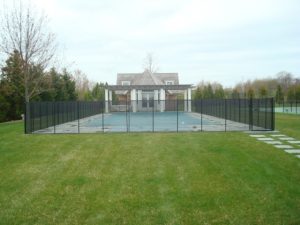 Black mesh pool fence surrounds a covered pool with a house in the background and a green lawn in the foreground on Long Island.