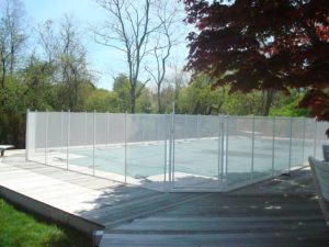 White mesh pool fence encloses a covered pool, set on a wooden deck surrounded by lush trees and a red-leaved tree in the foreground.