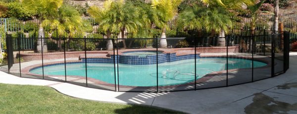 A swimming pool in hampton enclosed by a pool safety fence, featuring a tub connected to the pool and is surrounded by trees, and neighboring house in the background.