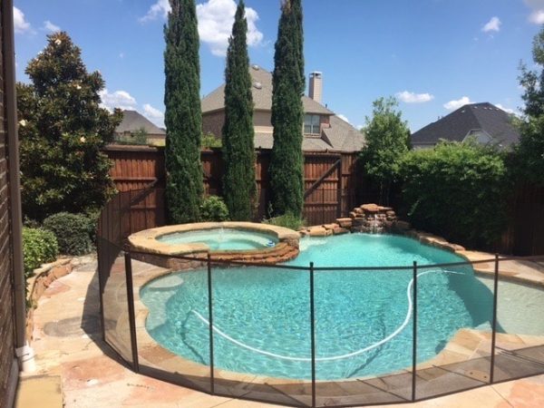 A swimming pool in land o lakes enclosed by a pool safety fence, featuring a hot tub connected to the pool and is surrounded by tall trees, a wooden fence, and neighboring house in the background under a clear sunny sky.