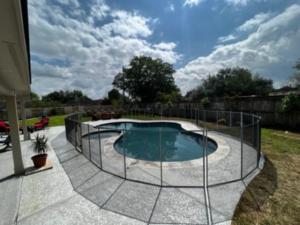 A swimming pool in paterson enclosed by a pool safety fence on a large, paved patio and is surrounded by a wooden privacy fence, and red lounge chairs.