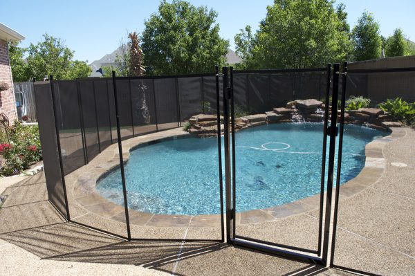 A pool in volusia enclosed by a black mesh pool fence with a secure gate surrounded by a stone border and trees lining the yard.