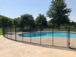 A curved swimming pool in atlanta with a black mesh safety fence placed around its border, set against a backdrop of neatly trimmed lawn and large trees. The secure fencing offers a clear view of the pool and surrounding nature, emphasizing safety without obstructing the scenery.