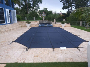 Pool cover secured over a rectangular pool on a stone patio, with tropical plants and a modern house in long island