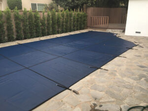 Blue safety pool cover secured over a rectangular pool on a stone patio in orange county, with a wooden deck and a row of tall shrubs in the background.