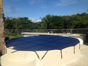 Blue mesh pool cover secured over a circular pool with a beige raised edge in orlando, surrounded by trees and a chain-link fence.