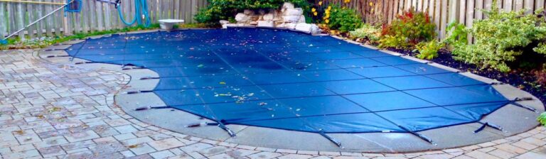 A backyard pool in atlanta covered with a blue safety pool cover, secured with metal clips around its edges. The surrounding area includes a stone-paved patio and a landscaped garden, with bushes and plants near a wooden fence.