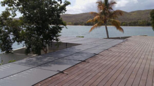 Mesh pool cover secured over an L-shaped pool in redlands beside a wooden deck, with a palm tree and waterfront view in the background.