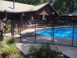 A curved swimming pool in palm bay fl with a black mesh safety fence placed around its border, set against a backdrop of neatly trimmed lawn and large trees. The secure fencing offers a clear view of the pool and surrounding nature, emphasizing safety without obstructing the scenery.
