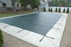 Green mesh pool cover tightly secured over a rectangular pool in a backyard with a wooden deck, patio furniture, and surrounding trees in tampa.