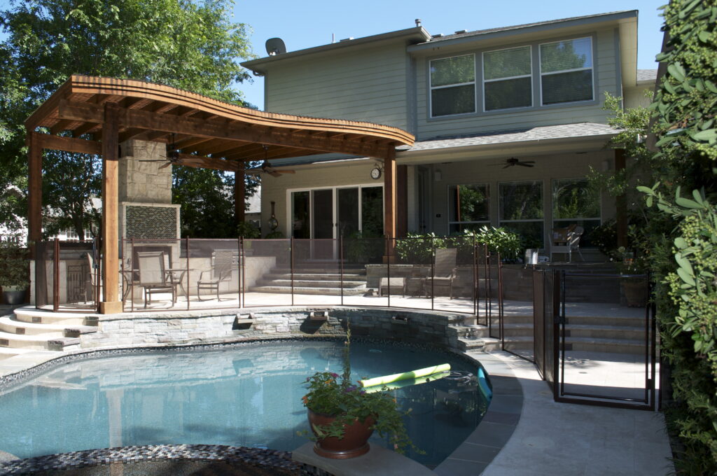 Modern backyard with a mesh pool fence enclosing a curved swimming pool, outdoor seating area, and wooden pergola-covered patio